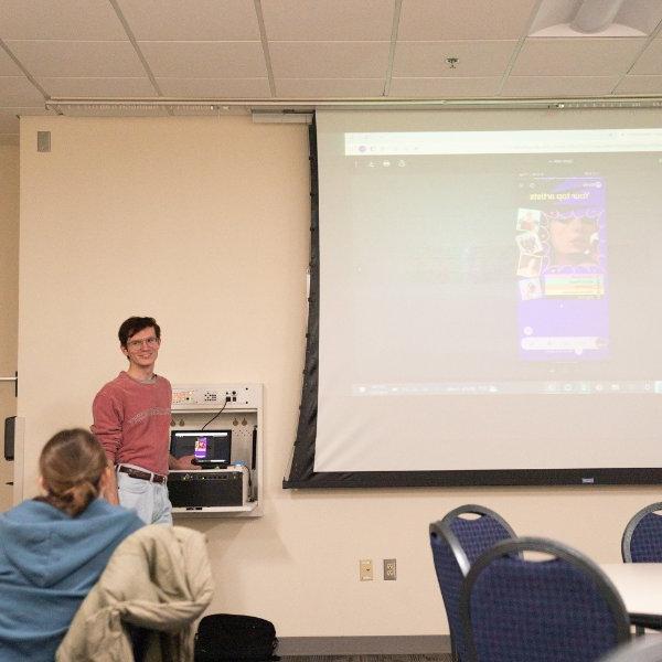 Caleb White站在投影屏幕的左边，屏幕上显示着Spotify上Taylor Swift的截图.
