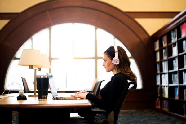  A college student wearing pick headphones works on a laptop in a university library. 